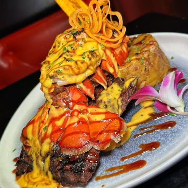Grilled steak and lobster tails topped with yellow sauce, garnished with fried onions and a pink orchid.