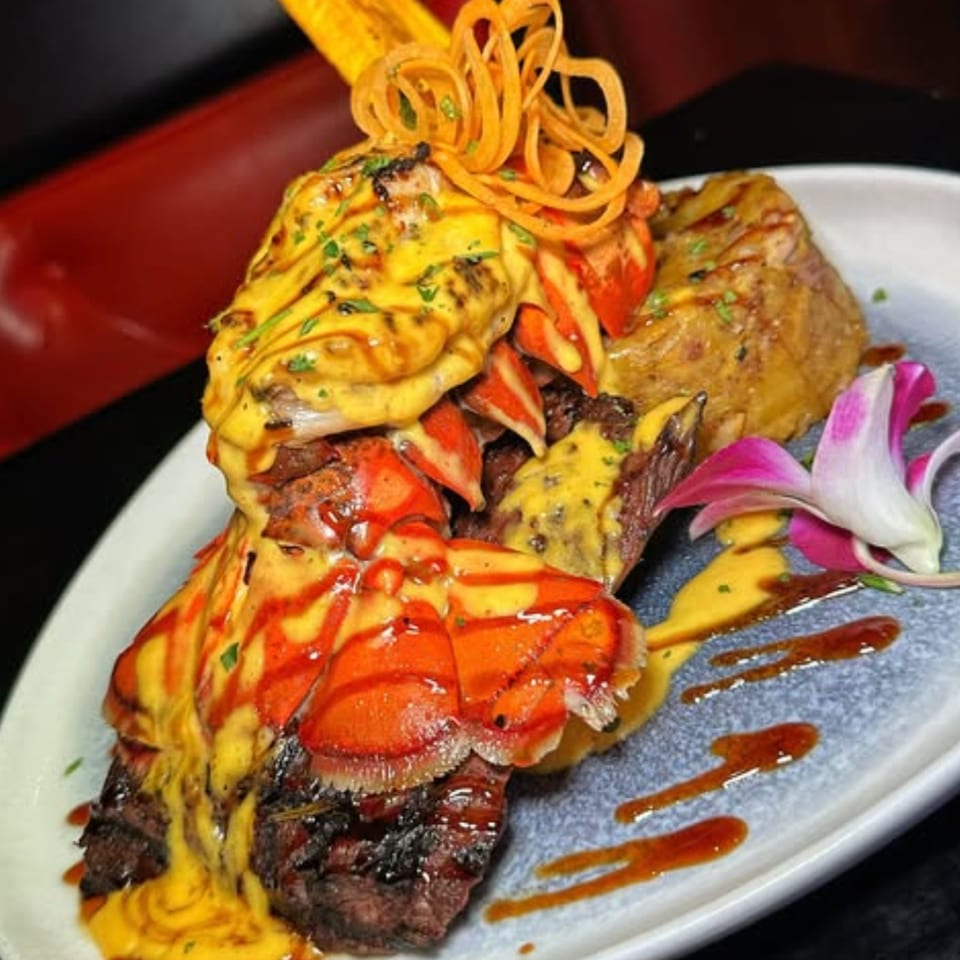 Grilled steak and lobster tails topped with yellow sauce, garnished with fried onions and a pink orchid.