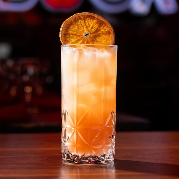 A tall, faceted glass filled with a light orange cocktail, ice, and a dried orange slice.