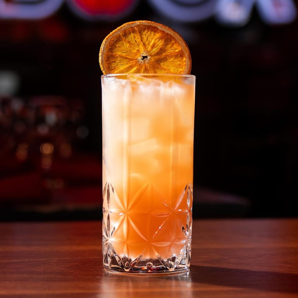 A tall, faceted glass filled with a light orange cocktail, ice, and a dried orange slice.