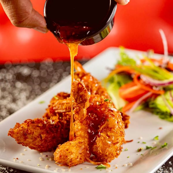 A hand pours a rich, golden sauce over crispy fried shrimp, served with a fresh side salad.