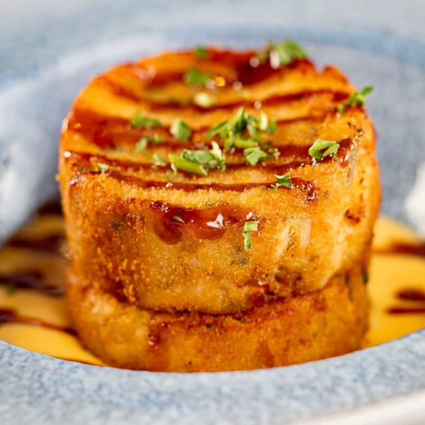 Golden-brown Latin-American potato cake, drizzled with sauce and garnished with fresh herbs, in a blue bowl.