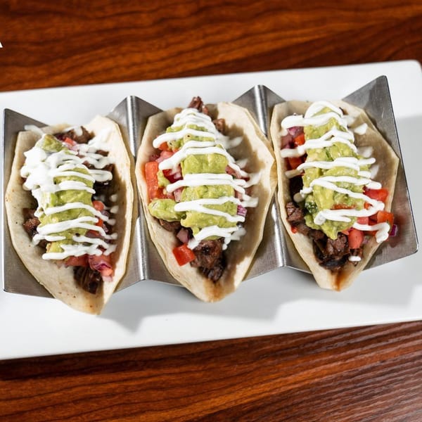 Three Latin-American tacos with meat, diced tomatoes, guacamole, and crema on a white plate.