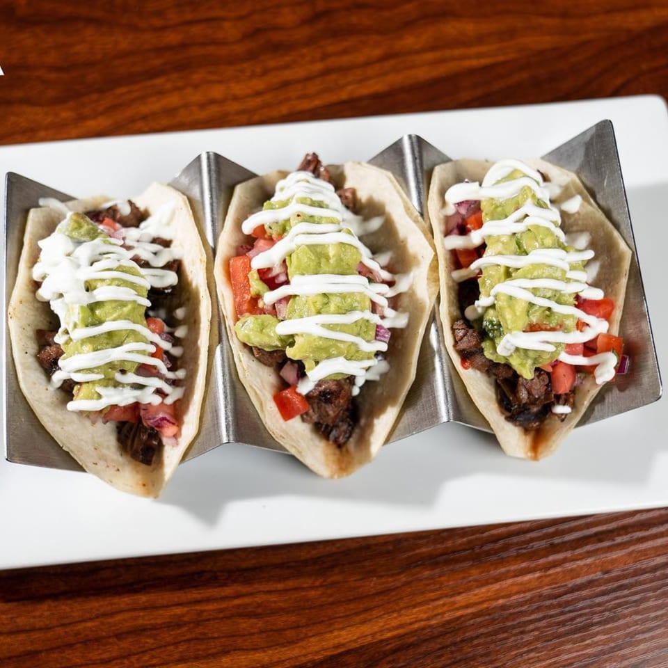 Three Latin-American tacos with meat, diced tomatoes, guacamole, and crema on a white plate.
