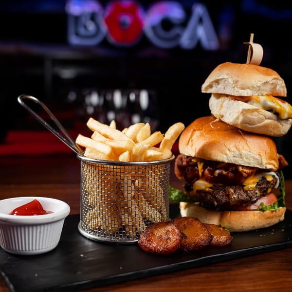 A large burger, a slider, fries in a metal basket, and plantains on a black tray.