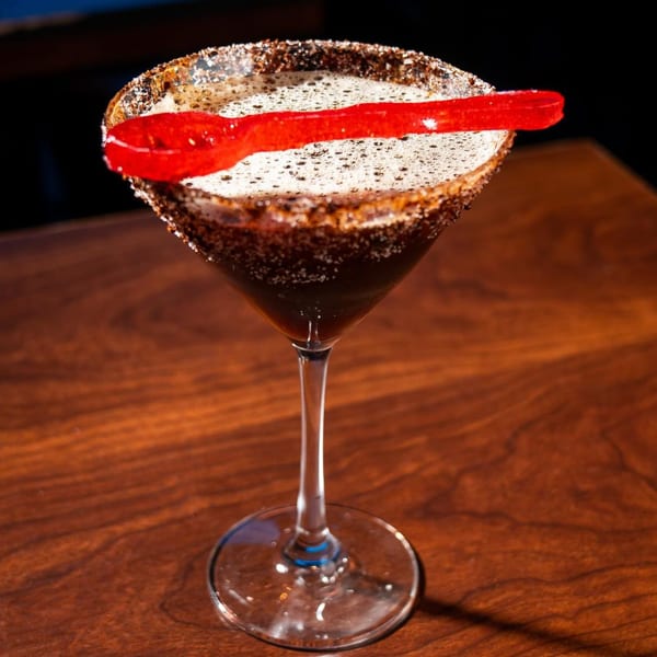 Dark cocktail in a martini glass with a sugar-rimmed edge and a red gummy candy garnish.