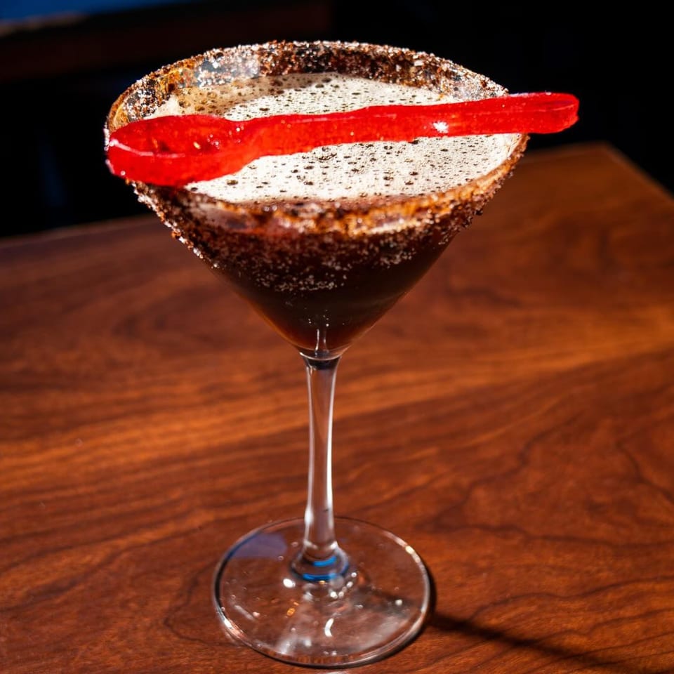 Dark cocktail in a martini glass with a sugar-rimmed edge and a red gummy candy garnish.