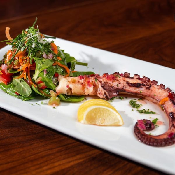Grilled octopus tentacle with a side salad and a lemon wedge on a white rectangular plate.