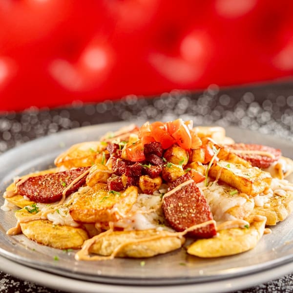 Close-up of a Latin-American dish with plantains, chorizo, cheese, and diced tomatoes on a metal plate.