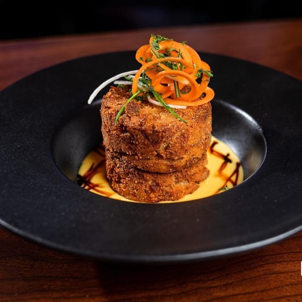 Crispy Latin-American croquettes stacked on a black plate with yellow sauce, garnished with carrot curls and herbs.