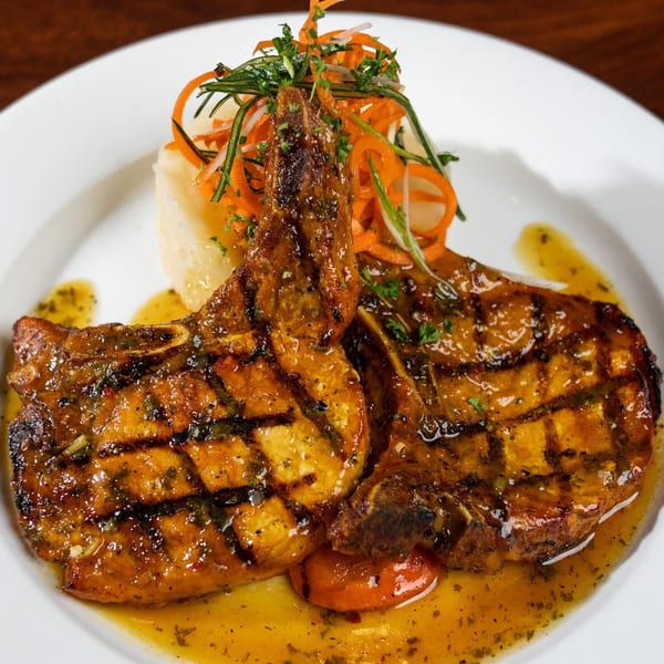 Two grilled pork chops with sauce, mashed potatoes, and julienned vegetables on a white plate.