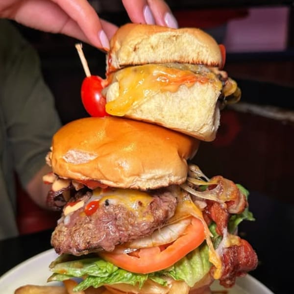 A hand holds a skewer through a towering stack of two Boca Restaurant burgers with cheese, bacon, and vegetables.