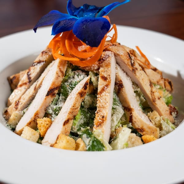 Grilled chicken Caesar salad with croutons, parmesan, carrot ribbons, and a blue orchid garnish.