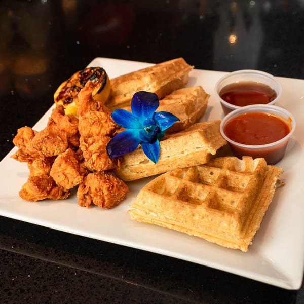 Chicken and waffles with a grilled lemon, two sauces, and a blue orchid on a white plate.